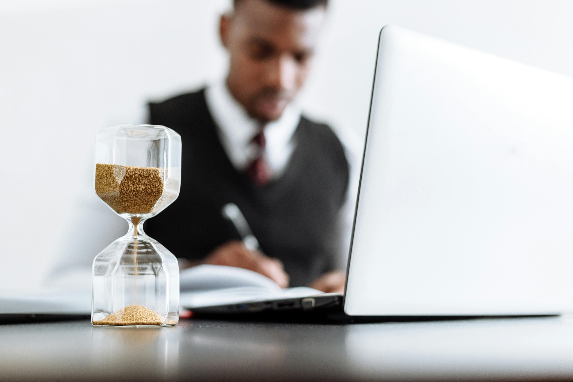 Hourglass on a desk in front of a businessman writing at a laptop