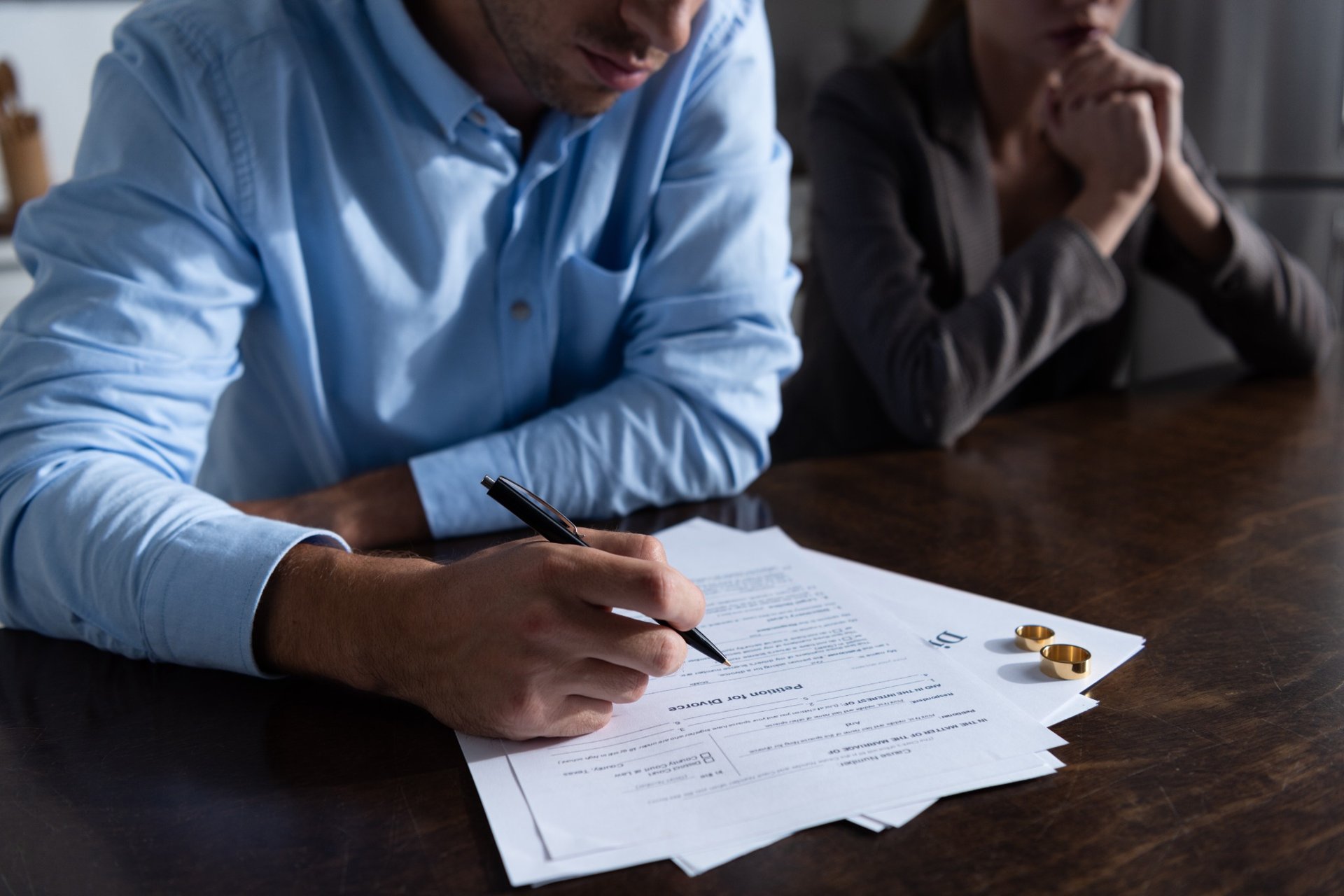 Man signing divorce papers while woman sits beside him at table