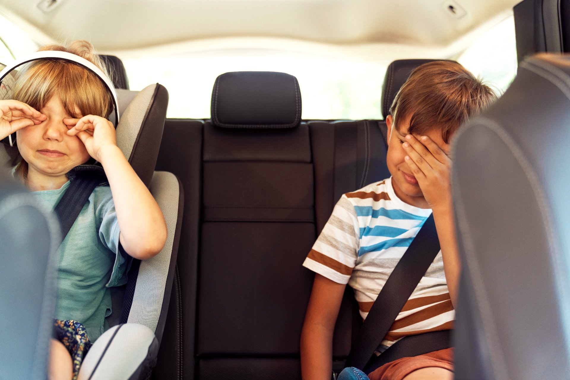 Two children in the backseat of a car rubbing their eyes while seated with seatbelts on