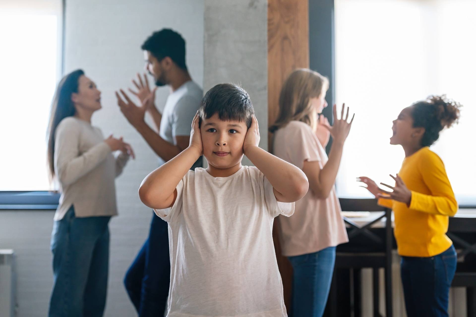 Sad boy covers his ears while adults and children argue in the background during a family conflict