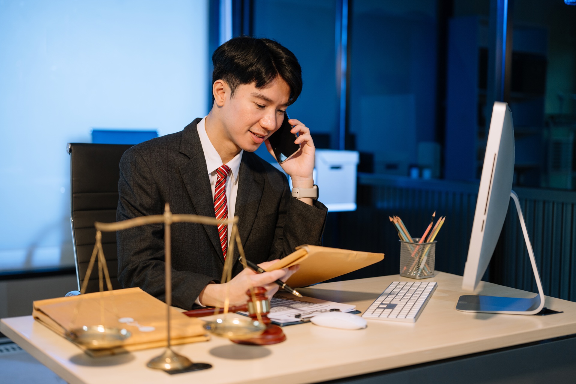 Male lawyer in suit speaking on phone at office desk at night, holding case file with justice scales and gavel on desk