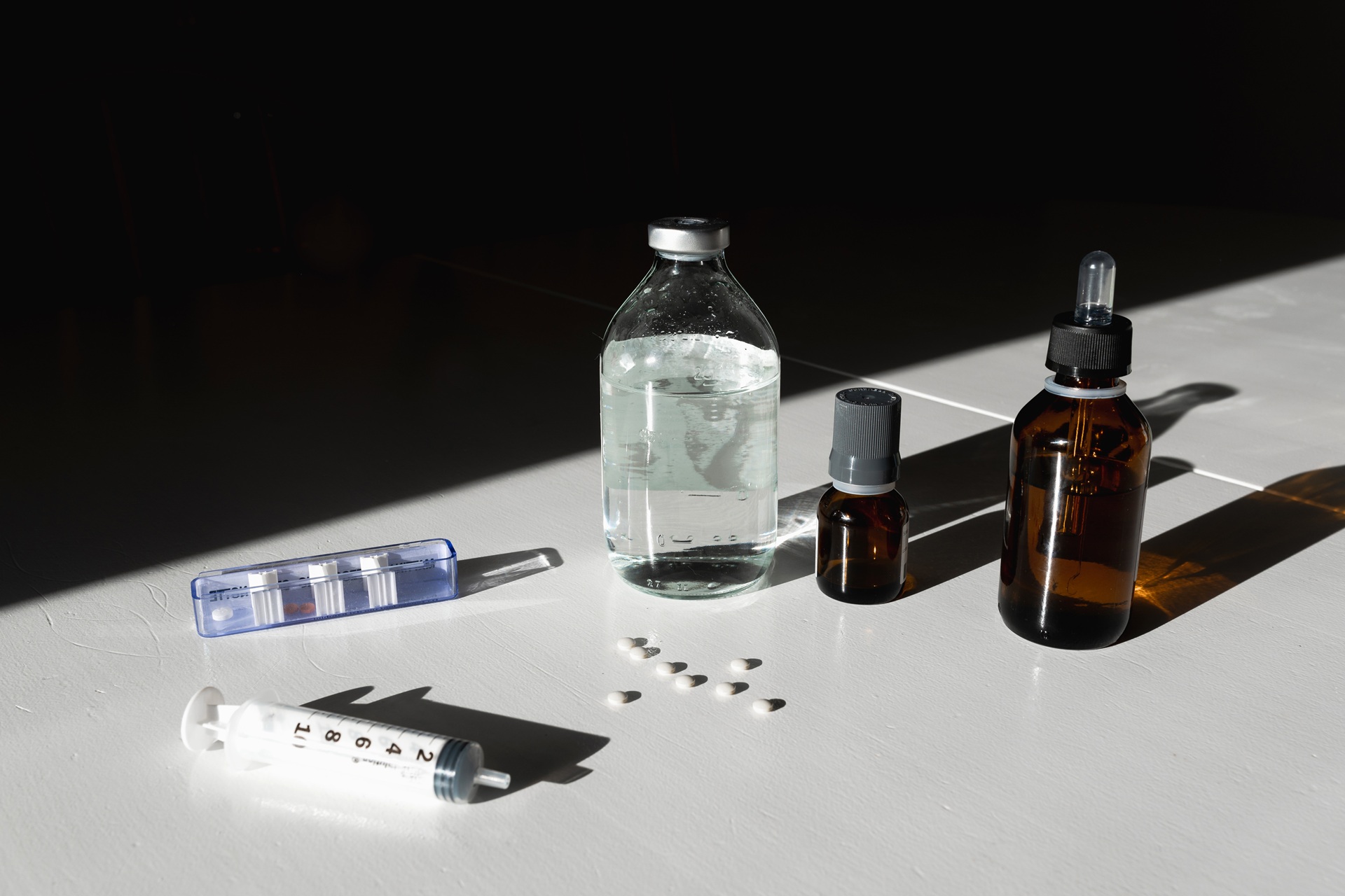 Medical supplies including a syringe, assorted medicine bottles, and scattered pills on a white table with strong shadows