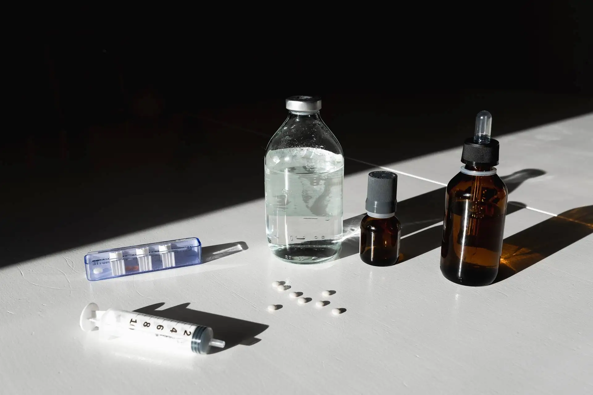 Medical supplies including a syringe, assorted medicine bottles, and scattered pills on a white table with strong shadows