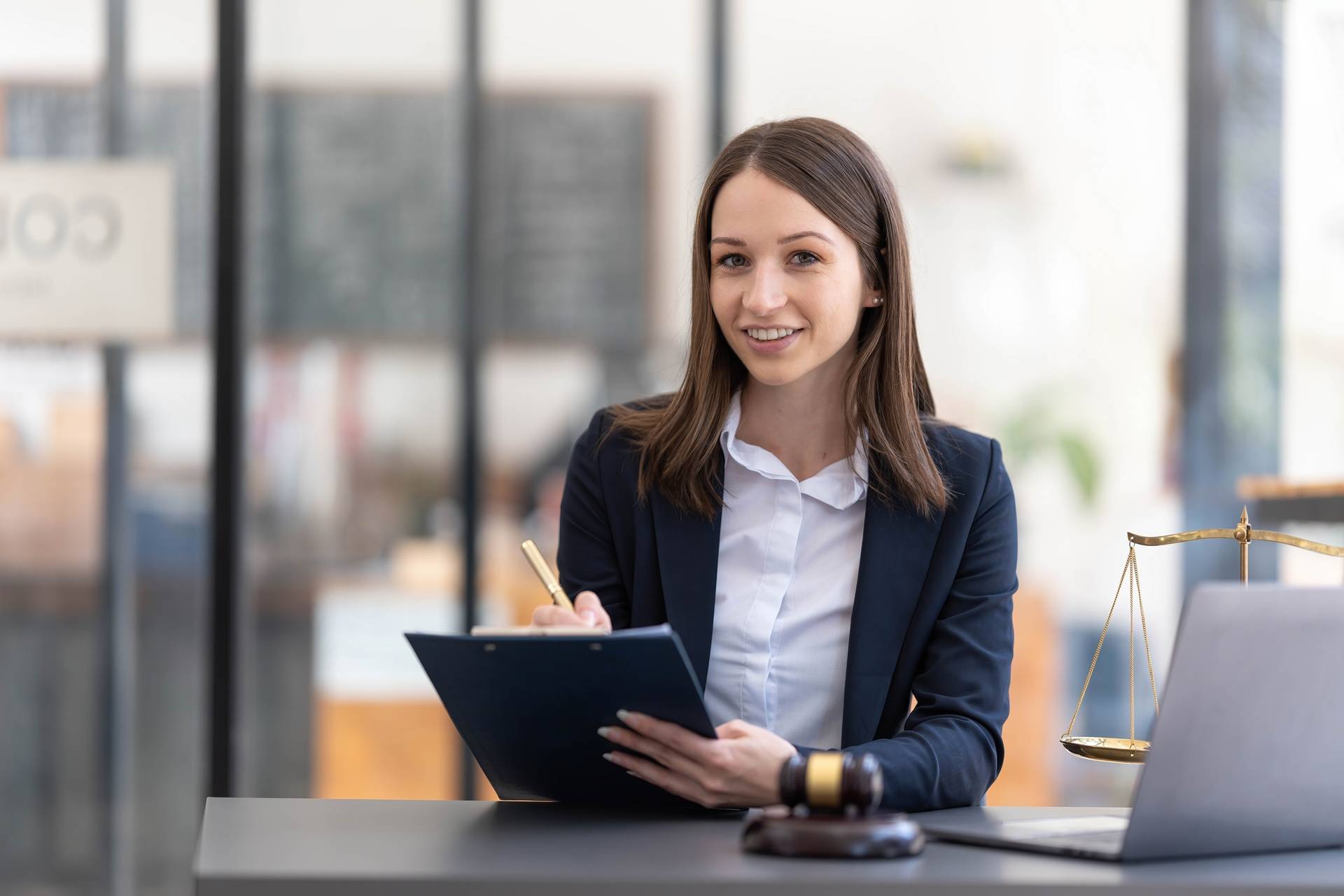A female lawyer in a navy suit smiles while holding a clipboard and pen at her desk, with a gavel, scales of justice, and a laptop in front of her