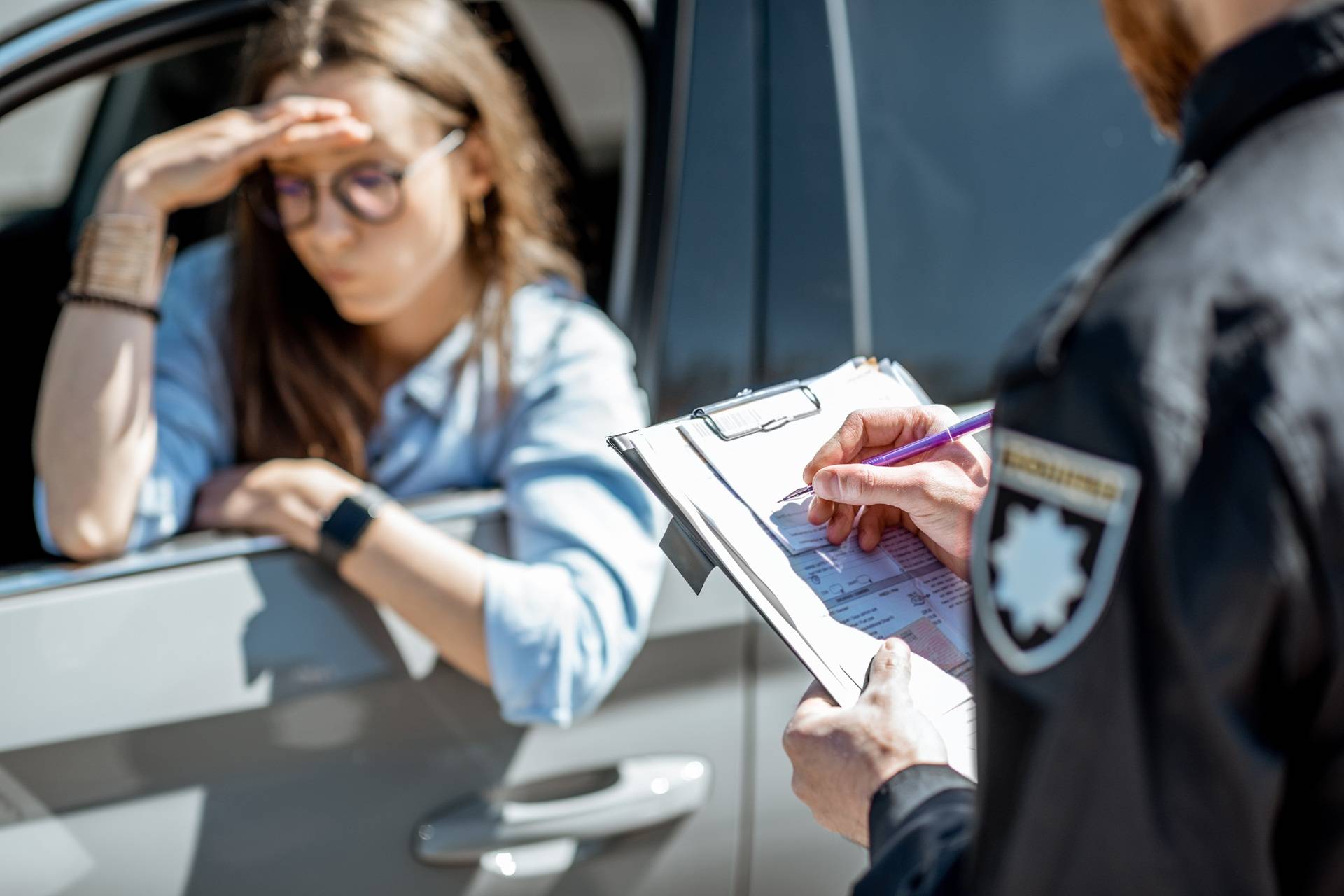 A police officer writes on a clipboard while a concerned woman in glasses leans on the window of her car, appearing stressed or frustrated