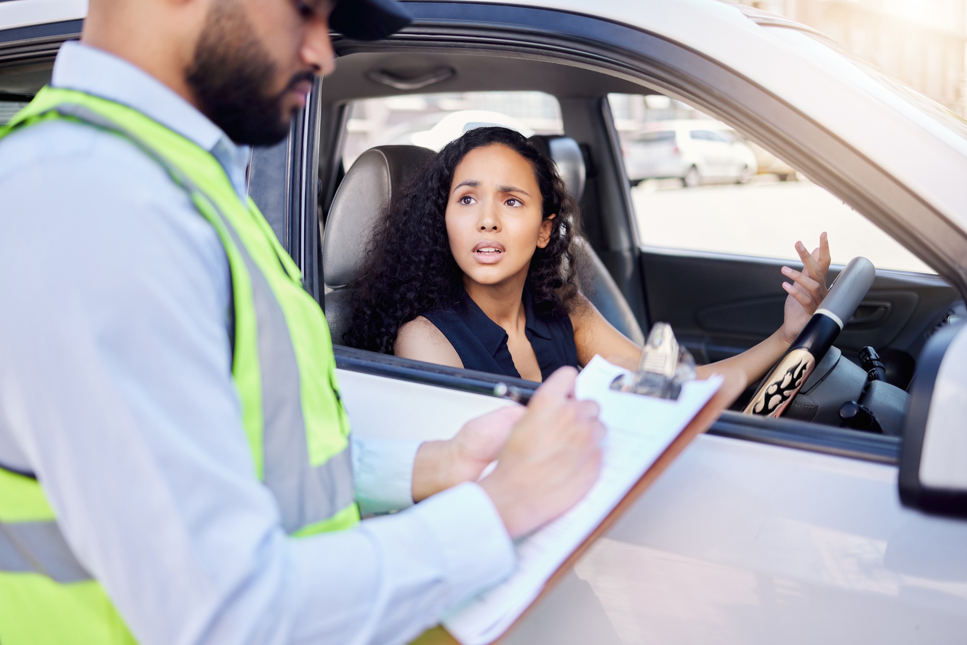 A young woman with curly hair sits in her car looking confused or upset, gesturing toward a male officer writing on a clipboard outside the vehicle