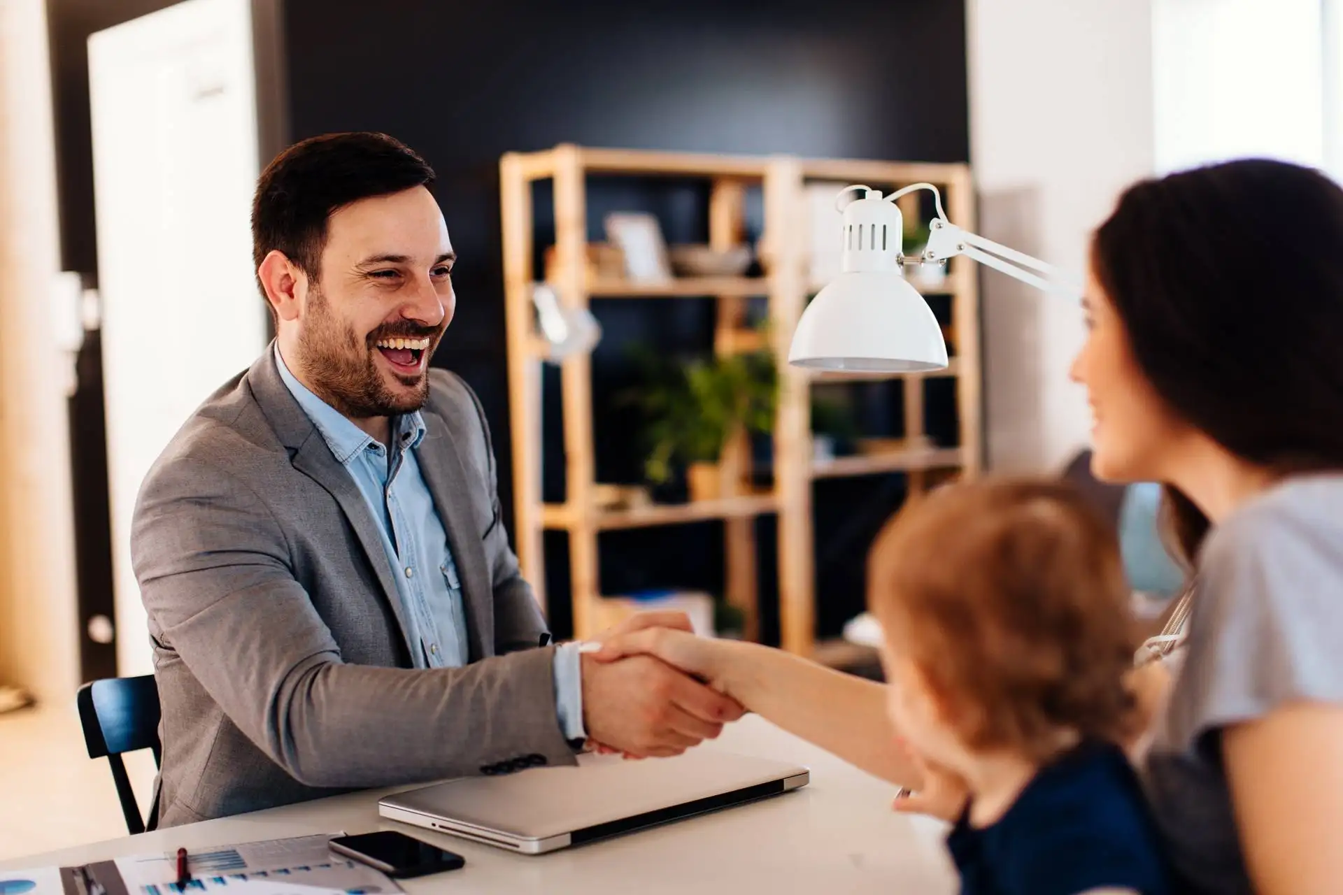 Smiling lawyer in a suit shaking hands with a woman across a desk, with a young child in her lap, suggesting a positive meeting or agreement