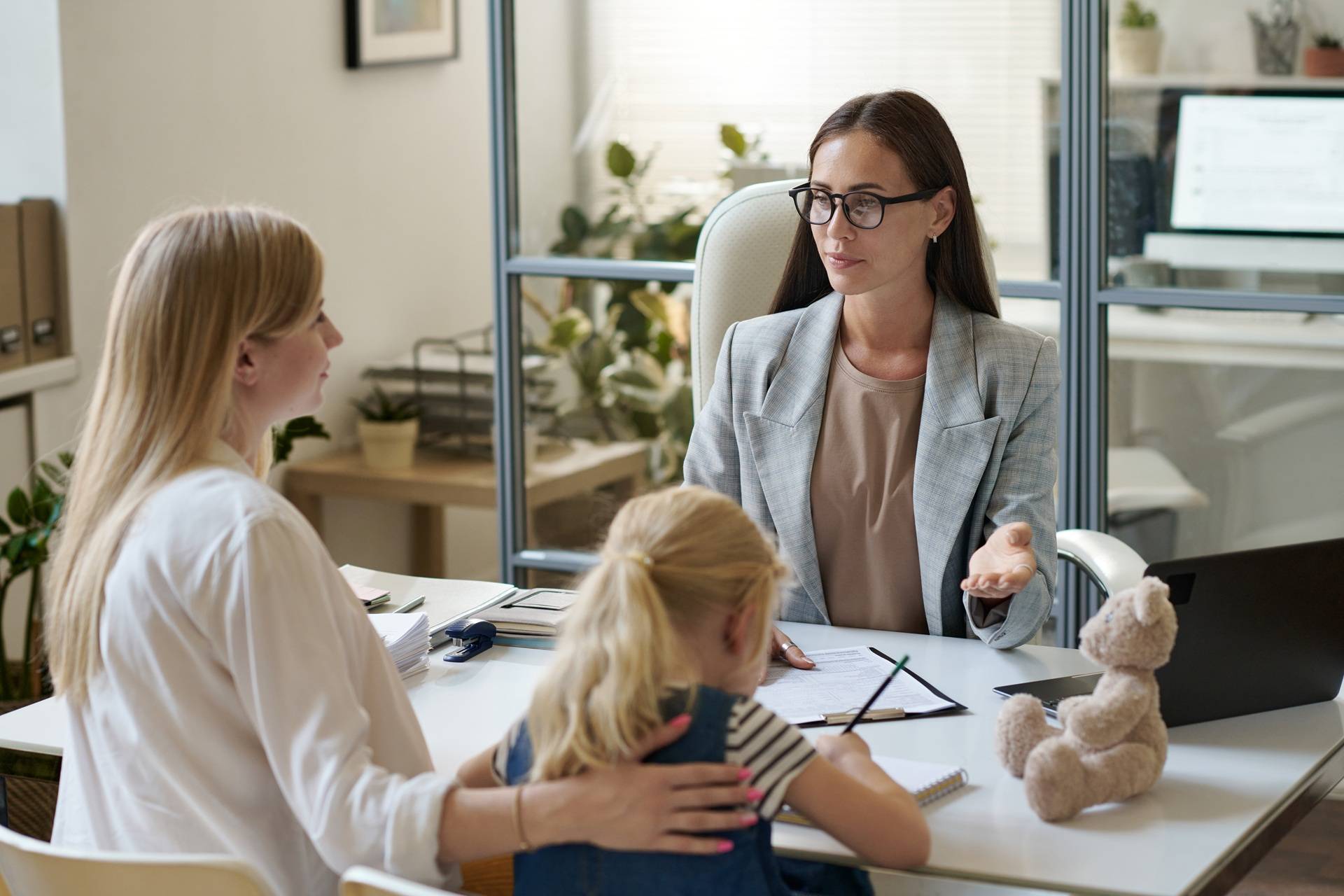 Female lawyer in an office setting talking to a woman and child across the desk, with the child drawing and a teddy bear on the table, indicating a legal or social consultation involving family matters