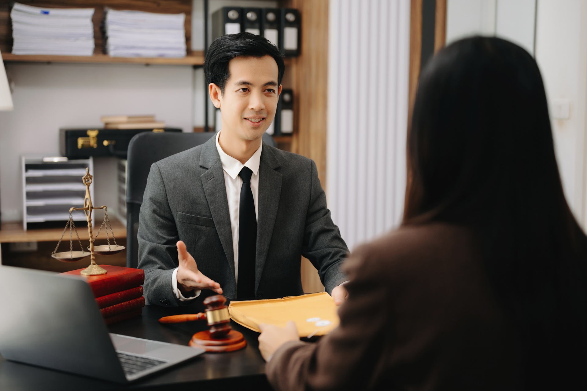 A male lawyer in a gray suit sits at a desk with legal documents, a laptop, a gavel, and justice scales, speaking to a client across the table