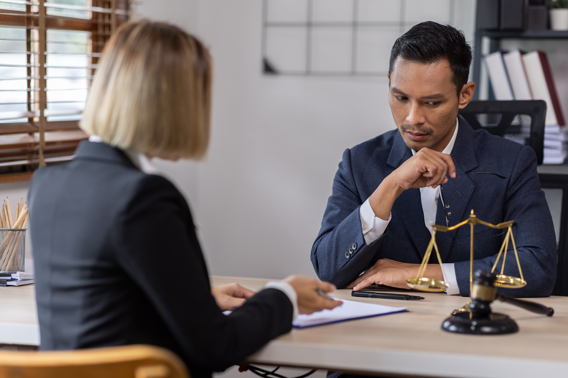 A serious-looking man in a suit sits at a desk with legal scales and a gavel, listening to a woman across from him holding a clipboard