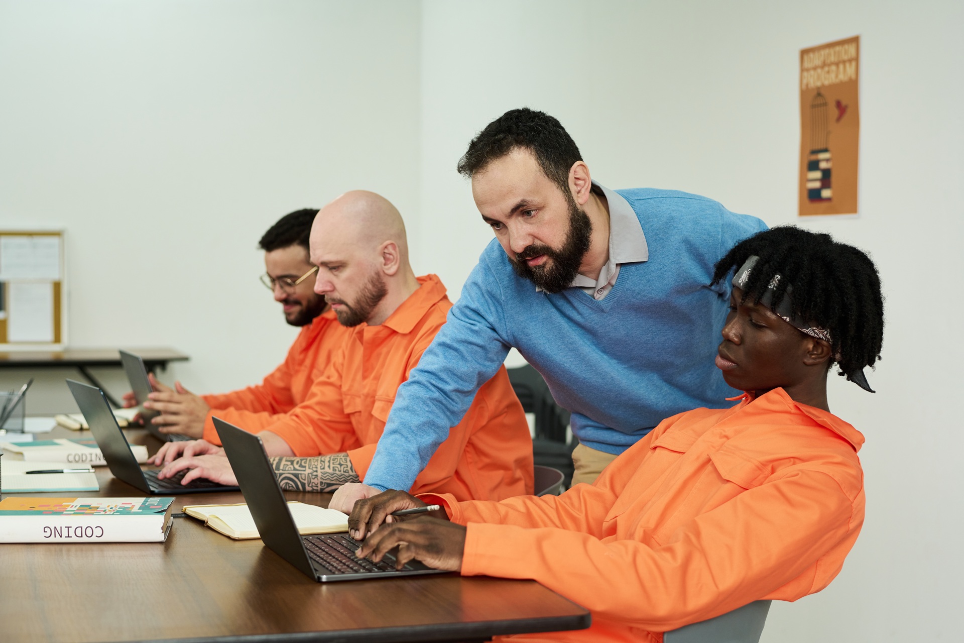 Three incarcerated men in orange uniforms work on laptops while an instructor in a blue sweater assists one of them in a classroom setting