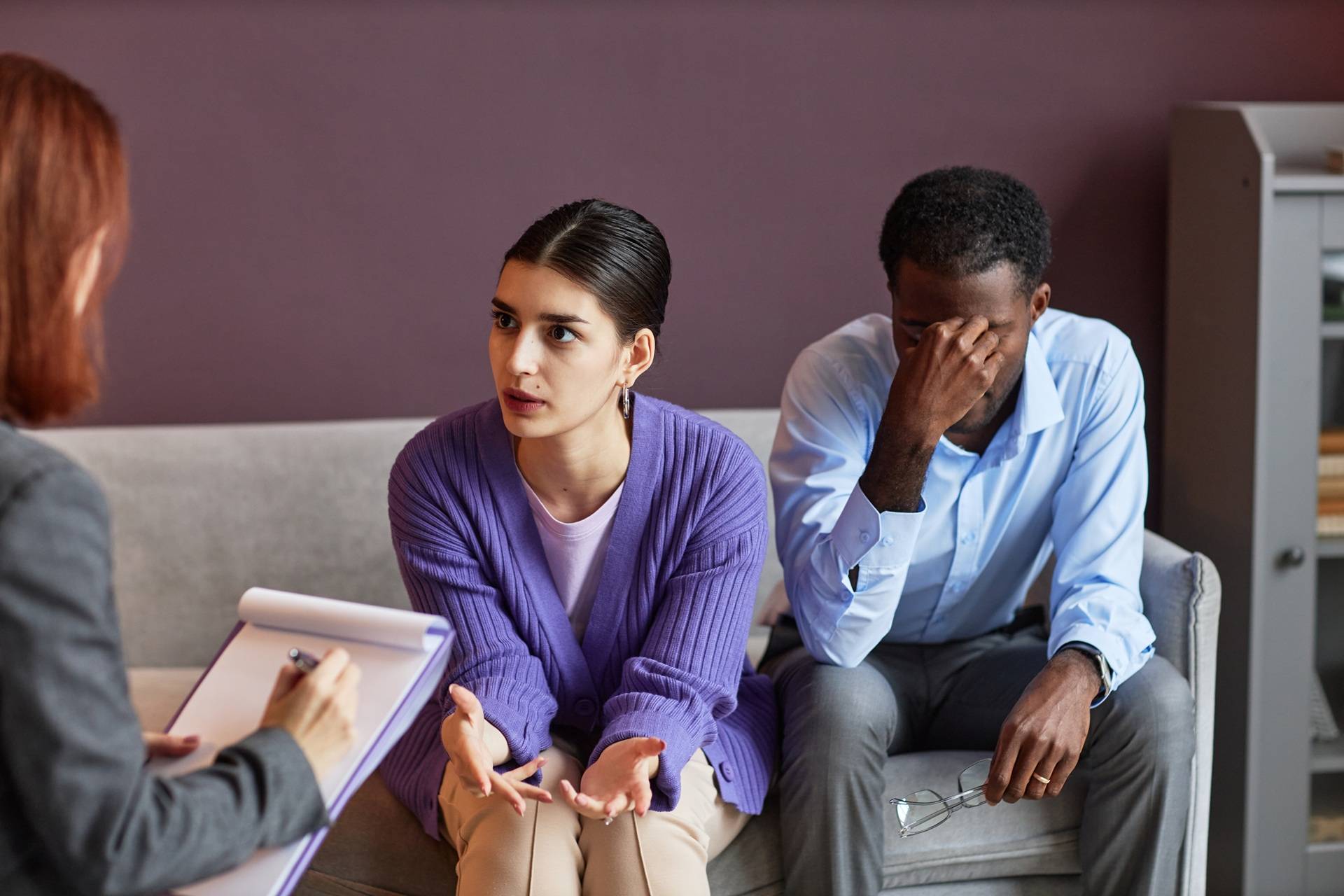 Couple in counseling session, woman talking emotionally while man sits beside her with hand on his face, therapist taking notes