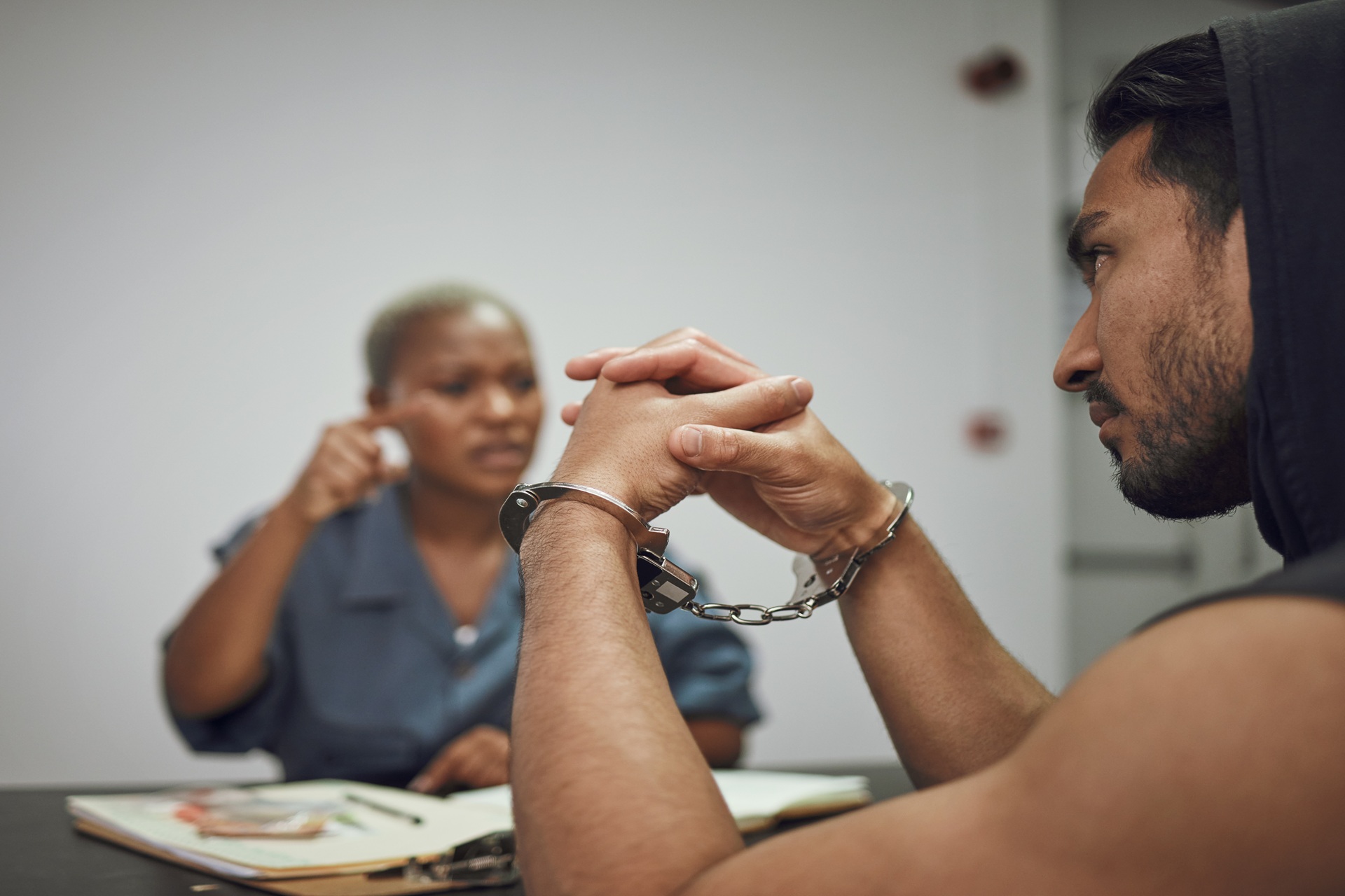 A man in handcuffs sits across from a woman speaking to him, with documents and a pen on the table between them