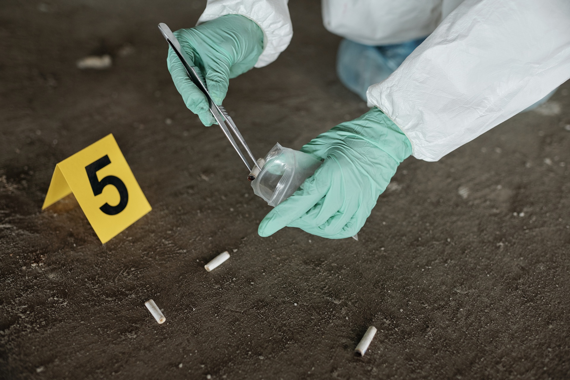 A forensic investigator wearing a white protective suit and green gloves uses tweezers to place a cigarette butt into an evidence bag near a yellow evidence marker labeled "5" on a concrete surface