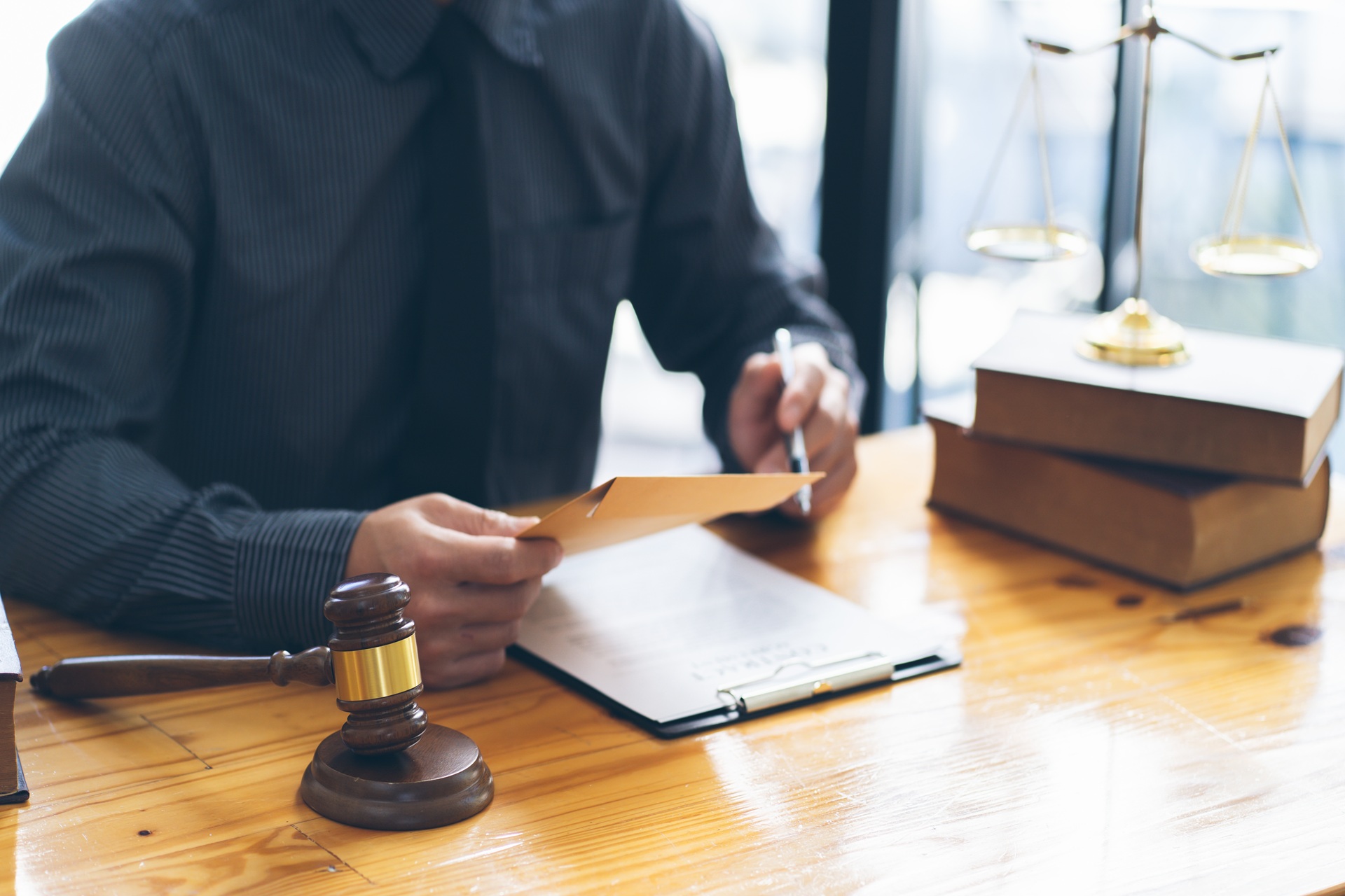 A lawyer in a dark shirt sits at a desk with legal books, a gavel, and a balance scale, holding an envelope and writing on a clipboard