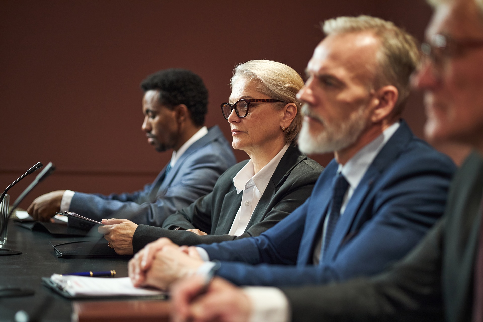 A panel of four attorneys in formal attire sits at a long table with microphones, listening attentively during the trial