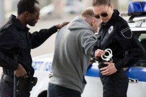 Two uniformed police officers detain a man near a patrol car, with one officer preparing to use handcuffs while the other stands close for support