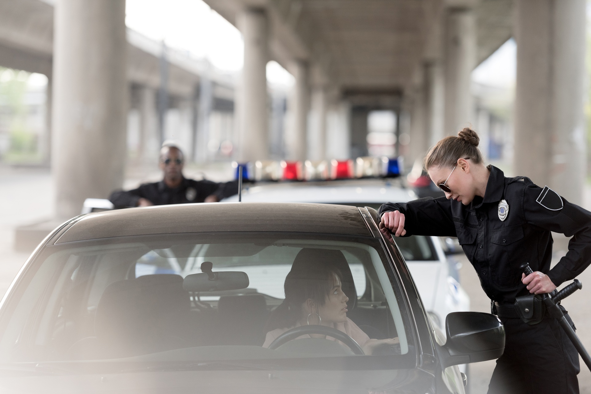 A policewoman in sunglasses leans on a car window speaking to a female driver, while another officer stands near a patrol car in the background