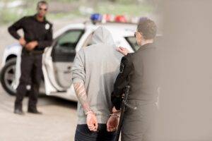 A female police officer in uniform placing her hand on the back of a handcuffed male suspect wearing a gray hoodie, with a police car and another officer in the background