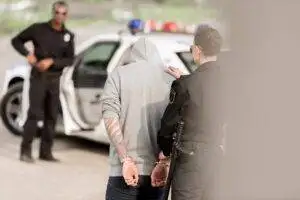 A female police officer in uniform placing her hand on the back of a handcuffed male suspect wearing a gray hoodie, with a police car and another officer in the background