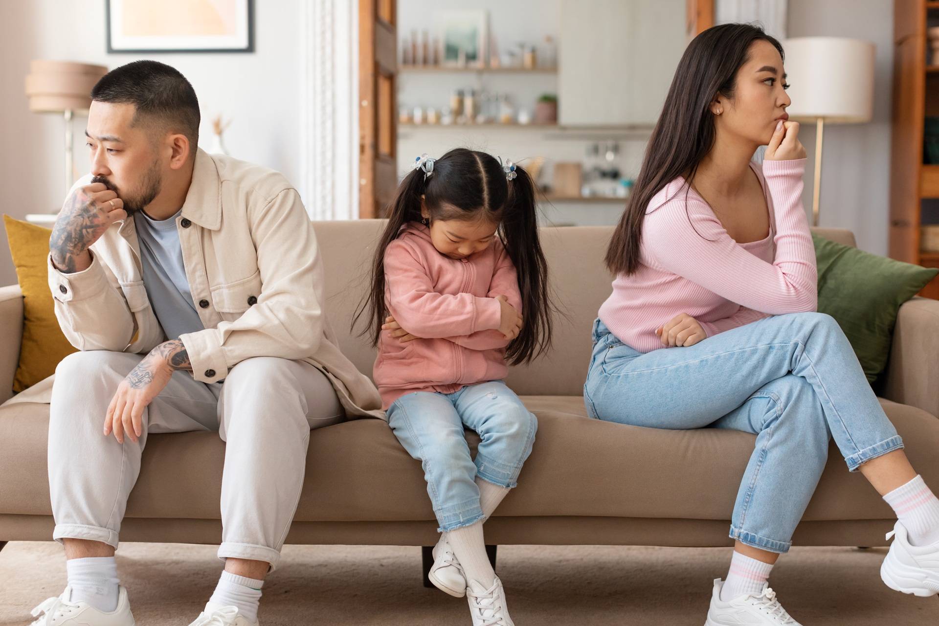 Young family in conflict sitting on sofa, child with head down between parents who are turned away from each other in silence